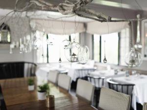 A casual fine dining area with tables covered in white tablecloths, black chairs, and glass ornaments hanging from white branches suspended from the ceiling, all bathed in natural light from large windows.