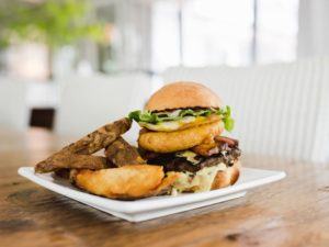 A Casual Fine Dining twist: A cheeseburger topped with lettuce, onion rings, and sauce is served on a square white plate with thick-cut potato wedges, set on a wooden table in a bright, relaxed space.