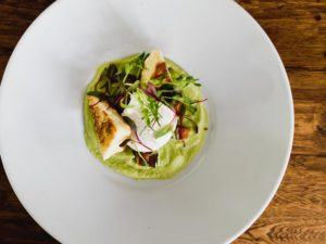 A white plate with a portion of grilled chicken, microgreens, and a poached egg on a bed of green puree, set on a wooden table—perfect for enjoying casual fine dining in an inviting atmosphere.