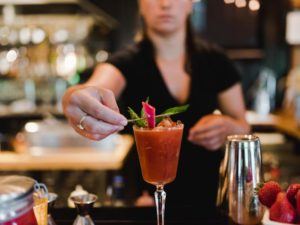 A bartender in a casual fine dining setting garnishes a reddish cocktail in a glass with a green leaf and pink decoration, surrounded by bar tools and fresh strawberries on the counter.