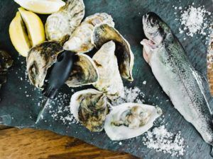 A whole raw fish, several oysters—some open, some closed—a black-handled oyster knife, and lemon wedges arranged on a dark slate with scattered coarse salt evoke the fresh elegance of casual fine dining.