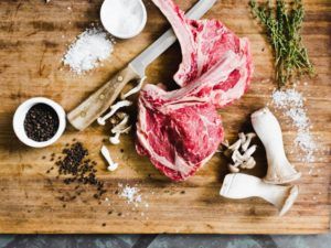 Two raw ribeye steaks on a wooden cutting board with a knife, fresh herbs, salt, peppercorns, and king oyster mushrooms create the perfect scene for casual fine dining at home.