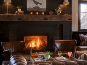 Cozy living room with a lit fireplace, two brown leather chairs, a rustic wooden mantel with candles and plants, and a tray with drinks and snacks on an ottoman. Warm lighting and optional concierge services create an inviting atmosphere.