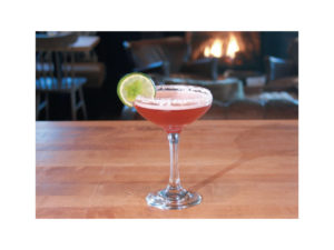A pink au currant cocktail in a stemmed glass with a salted rim and lime wheel garnish sits on a wooden table; a cozy fireplace and chairs are blurred in the background.
