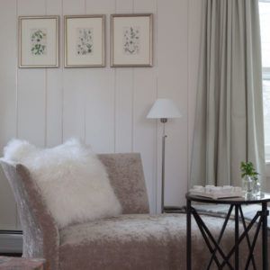 A cozy living room corner with a plush chaise lounge, a white fluffy pillow, a round side table topped with a plant and tray, a floor lamp, light curtains, and three framed botanical prints on a white paneled wall—so it goes.
