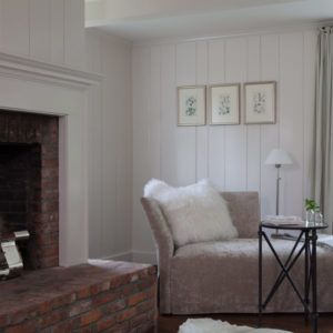 A cozy living room corner with a brick fireplace, a soft gray chaise lounge with a white fluffy pillow, a small round side table, and three botanical prints on a white paneled wall—so it goes, inviting warmth and tranquility.