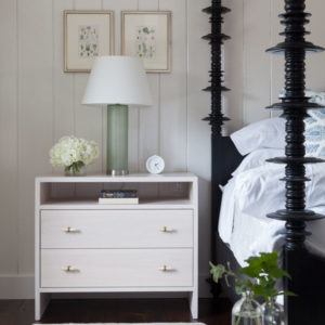 A white nightstand with two drawers holds a green glass lamp with a white shade, a small white alarm clock, and a vase of white flowers beside a tall black bedframe—so it goes in this cozy bedroom.