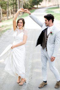 A bride and groom dance joyfully on a rural road. The bride lifts her dress slightly and smiles, while the groom twirls her, both looking happy and dressed in formal wedding attire. Trees line the background.
