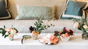 A white coffee table with small floral arrangements, a magnifying glass, and scattered flower petals sits in front of a beige sofa with decorative pillows in the background.