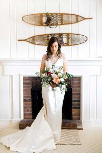 A bride in a white gown holds a large bouquet of flowers and stands in front of a brick fireplace. Two vintage snowshoes are mounted on the white wall above her.