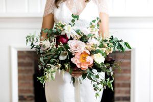 A bride in a white dress holds a large bouquet of flowers featuring pink, peach, and white blooms, green foliage, and delicate accents, with a blurred fireplace background.