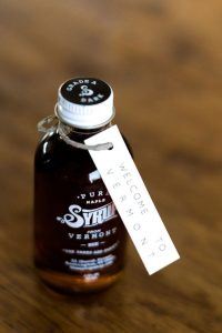 A small glass bottle of pure maple syrup from Vermont sits on a wooden surface, with a white tag attached that reads Welcome to Vermont.