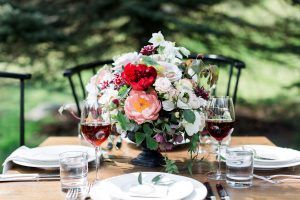 A wooden table set for two with wine glasses, plates, cutlery, water glasses, and a large, colorful floral centerpiece. Two black chairs are visible, and trees are blurred in the background.
