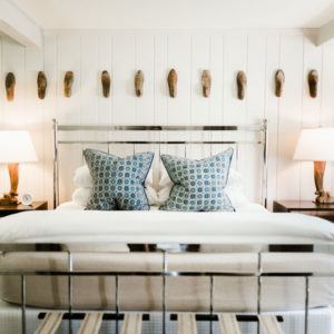 A neatly made bed with a metal frame, two patterned blue and white pillows, and two wall-mounted lamps on bedside tables. Be Positive artwork and wooden decorative pieces are displayed on the white paneled wall above the headboard.