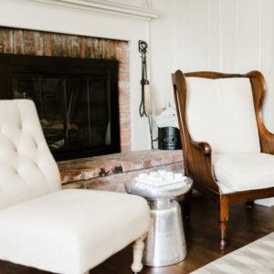 A cozy, inviting living room with a brick fireplace encourages you to be positive. Two cream-colored upholstered chairs and a metallic side table with a bowl on top create the perfect spot to relax by the fire, with fireplace tools beside the hearth.