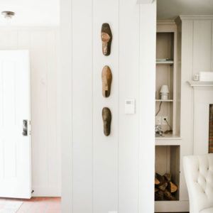 A white, sunlit room radiates a be positive vibe, with three wooden shoe molds mounted vertically on the wall. To the right, a built-in shelf and part of a fireplace appear, while an open door lets in uplifting natural light from the left.
