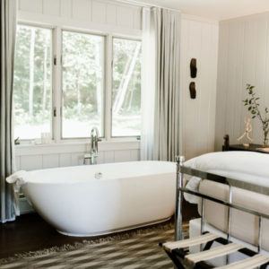 A bright, Be Positive bathroom features a freestanding white bathtub by large windows with gray curtains, next to a bed with white linens and metal frame, all atop a striped rug on a dark wooden floor.