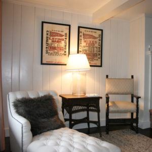 A cozy corner featuring a white tufted chaise lounge with a dark pillow, a wicker side table with a lamp, an upholstered chair, and two framed carpe diem posters on white paneled walls.