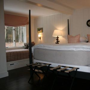 A cozy bedroom with a four-poster bed, neutral bedding, two pillows, and a wooden lamp on a nightstand. A window seat with cushions offers views of trees and fallen leaves—an inviting space to relax and embrace carpe diem.