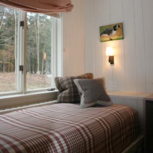 A cozy bedroom corner with a single bed covered in a plaid blanket, patterned pillows, and a wall lamp invites you to relax. A carpe diem print beside the bird painting inspires seizing the day as sunlight filters through the large window.