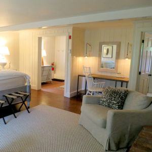 A cozy bedroom with a large bed, a desk with a mirror and chair, and a cushioned armchair with a patterned pillow invites you to relax. Soft lighting and wooden floors reflect the do unto others spirit in this welcoming space.