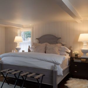 A cozy bedroom with a wooden bed topped with white bedding and beige pillows, flanked by two nightstands with large white lamps. The room’s calm ambiance gently echoes the peaceful do unto others spirit.