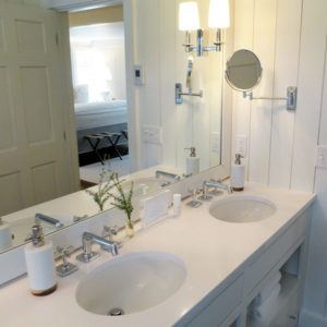 A modern bathroom with a double sink vanity, chrome faucets, wall-mounted mirror, white walls, lights, and a small plant—designed with a do unto others touch. A bedroom is visible through an open door in the background.