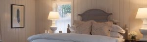 A cozy bedroom with wood-paneled walls, a wooden headboard, two white lamps on bedside tables, floral pillows, framed art—one featuring the phrase “do unto others”—and sunlight streaming through two small windows.
