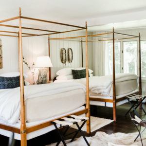 Two wooden four-poster beds with white bedding and black accent pillows are side by side in a bright bedroom, where white paneled walls, mirrors, and large windows let in natural light—creating a space where your dream sleep can be realized.