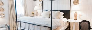A bright, cozy bedroom with a black metal canopy bed, white bedding, two pillows, bedside tables with owl lamps, wall art featuring If not now when, a window with beige curtains, and a small vase of yellow flowers.