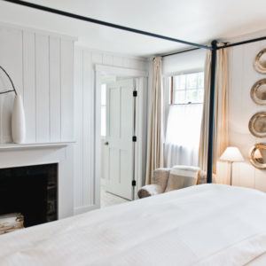 Bright, airy bedroom with white walls and linens. A cozy chair and lamp sit near a window, silver plates hang on the wall, and a vase decorates the mantel above the fireplace. An open door gently whispers: if not now when?.