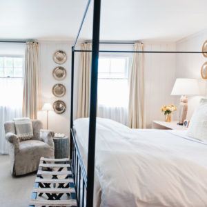 Bright bedroom with a four-poster bed, cream curtains, silver decorative plates, plush armchair, and fresh flowers on a white nightstand—an inviting space that whispers, “if not now when?”.