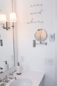 A modern bathroom vanity with a round sink, soap dispenser, and wall-mounted mirror features light fixtures and a playful wire sign that reads rise and shine above the thermostat and light switches.