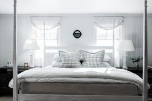 Bright, airy bedroom with a white four-poster bed, plaid pillows, and two white lamps on dark nightstands—perfect for rise and shine mornings with natural light streaming through the curtained windows behind the bed.