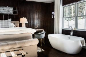 A cozy bedroom with dark wood walls, a beige upholstered bed, and a freestanding white bathtub by large windows. Show love with thoughtful touches like a lamp-lit nightstand, a small desk, and charming views of the snowy landscape outside.
