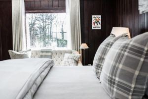 A cozy bedroom with a large bed in the foreground, plaid pillows, and a light sofa by a window. Dark wooden walls and soft natural light create a warm space to show love, highlighted by a lamp and ski-themed poster on the wall.