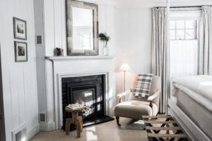 A cozy, bright bedroom where you can rise and shine, featuring white walls, a fireplace, a beige armchair with a plaid blanket, a wooden stool, a mirror above the mantel, and sunlight streaming through sheer curtains on the window.