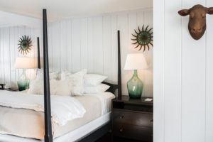 A cozy bedroom with white wood-paneled walls, a neatly made bed with white and beige bedding, green glass lamps on dark bedside tables, sunburst wall decor, and a mounted animal head inspires you to laugh everyday.