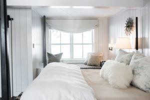 Cozy bedroom with white walls and bedding, decorative pillows, a soft throw blanket, and a “Laugh Everyday” sign above the bed. A window with a roman shade and a lamp on the nightstand add warmth to this cheerful retreat.