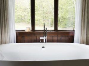 A white freestanding bathtub is positioned in front of a large window with white curtains, overlooking a green, leafy outdoor scene—perfect to let it be your tranquil escape. A modern faucet is mounted on a wooden panel beneath the window.