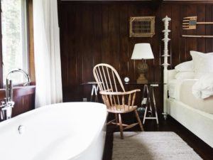 A cozy bedroom with dark wood walls, a white freestanding bathtub, and a bed with pillows invites you to relax and let it be; natural light pours in through a window with white curtains beside a wooden chair and glass desk.