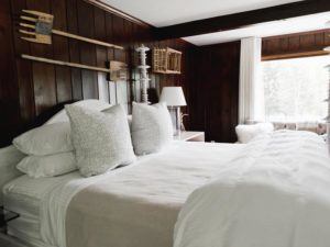 A cozy bedroom with a neatly made bed featuring white linens and large pillows, dark wood-paneled walls, modern lamps, and rustic wall decor; sunlight streams in through a large window with white curtains—just let it be your peaceful retreat.