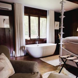 A modern white freestanding bathtub sits by a large window with light curtains in a cozy room with dark wood-paneled walls, hardwood floors, and soft furnishings—just let it be your perfect sanctuary for relaxation.