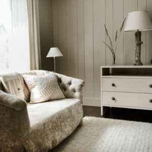 A cozy living room corner invites you to live in the moment with a plush beige loveseat, textured pillows, a white rug, and a light wood side table featuring two lamps and a simple vase of dried grasses against paneled walls.