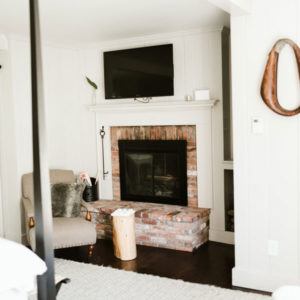 A cozy living room corner invites you to live in the moment with its brick fireplace, TV above, cushioned armchair and fur pillow, wooden stump side table, and a small stack of firewood.