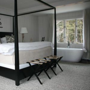 A modern bedroom with a black four-poster bed, white bedding, and two folding benches invites you to live in the moment. A freestanding bathtub by the window, light walls, and neutral decor create a serene retreat.