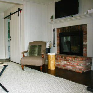 A cozy living room invites you to live in the moment, featuring a brick fireplace with a wall-mounted TV above, an upholstered armchair with a green pillow, a wooden stump side table, and a white sliding barn door.