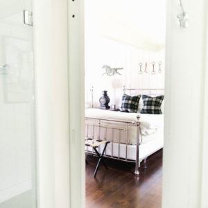 A bright bedroom with a metal bed frame, plaid pillows, dark wooden floor, and wall decor featuring a horse and keys invites you in—it’s now or never to experience this cozy space through the doorway.
