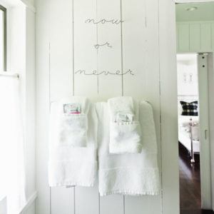 Two white towels hang on a white paneled wall, each with a washcloth and toiletries in the fold. Above the towels, wire art boldly spells “now or never” as natural light streams in from a window on the left.