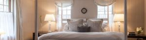 A cozy bedroom with a large bed, white and gray pillows, two bedside tables with lamps, and two windows with Roman shades on either side of the headboard invites you to rise and shine in soft lighting and tranquil, neutral colors.